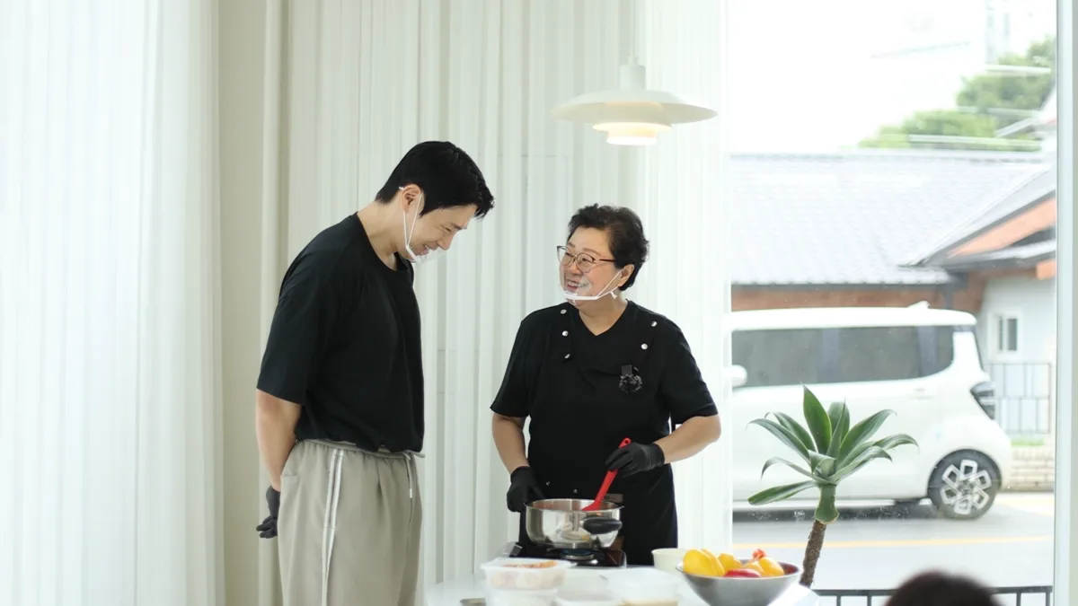 흰 커튼이 드리운 밝은 주방에서 검은 옷차림의 젊은 남자와 안경 쓴 여자가 마주 보고 웃는다. 여자가 냄비에 빨간 주걱을 넣고 젓는다