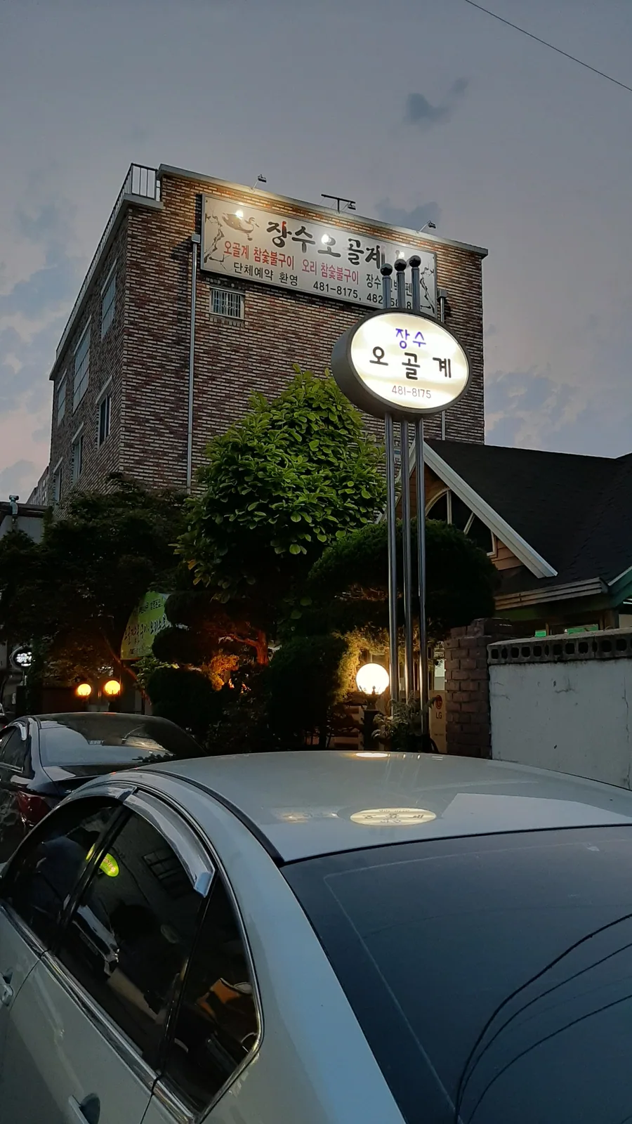 해 질 무렵 장수오골계숯불구이 건물 바깥 모습. 벽돌 건물 위에 장수오골계 간판이 켜져 있고 길가에 둥근 입간판이 서 있다.