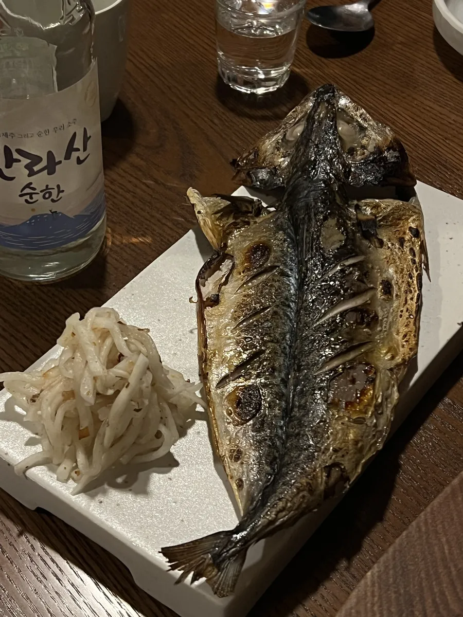 괸당 신당본점 고등어구이. 한 마리를 통째로 펼쳐 놓은 고등어가 흰 접시에 놓였고 옆에 무채와 한라산 순한 소주병이 함께 찍혀 있다.