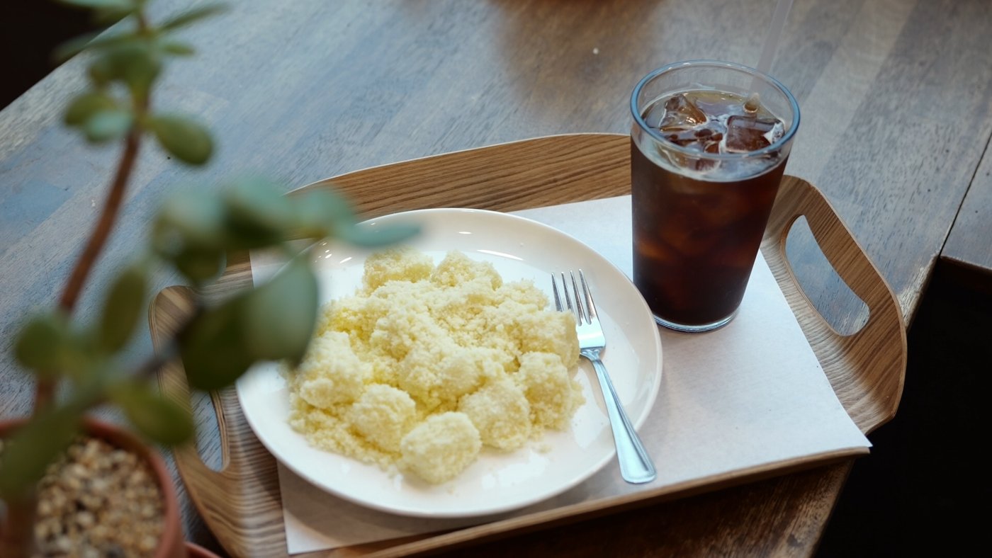 흰 접시에 노란 카스테라인절미 조각이 수북하고 포크와 아이스아메리카노가 나무 쟁반에 놓여 있다