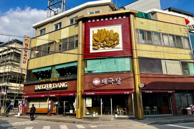 모서리 3층 건물, 금색 기둥, 붉은 간판 태극당과 TAEGEUKDANG Bakery & Cafe, 세로 간판 태극당과자점, 빵 그림 액자가 걸려 있다
